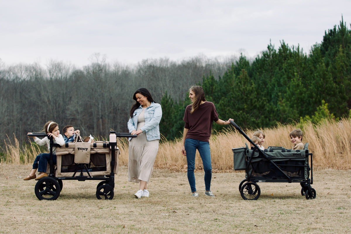 Two women pushing Keenz stroller wagons with children in a grassy field.