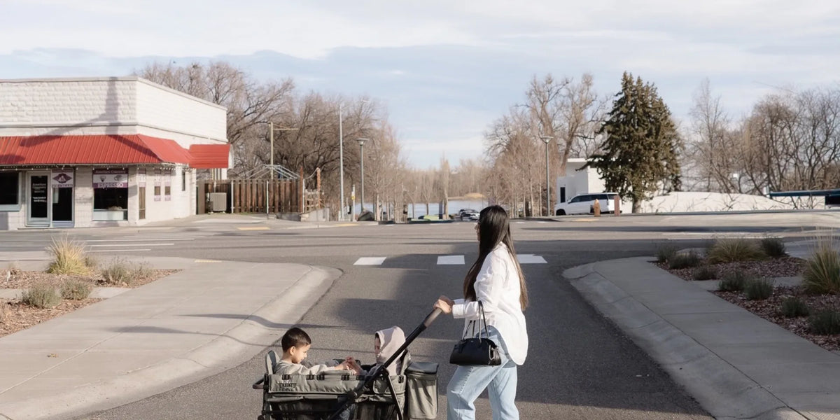 Mother pushing kids in a sage-green Keenz stroller wagon across a crosswalk