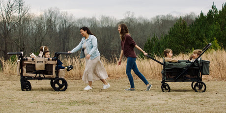A parent pushing a two-passenger Keenz MOOV 2 stroller wagon along a leafy park path with children seated inside.
